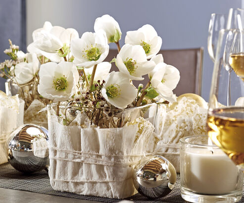 Christmas Rose as cut flowers in a glass wrapped in a waxed napkin, adorned with glitter balls and candles for a festive table decoration